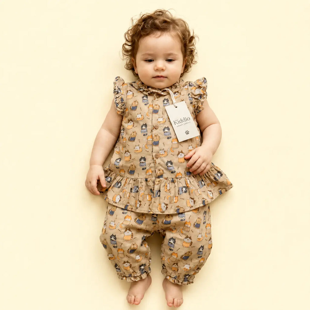 A toddler girl wearing a beige two-piece outfit with a cartoon animal print, featuring a ruffled sleeveless top and matching pants, lying on a solid pale yellow background.