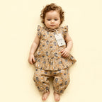 A toddler girl wearing a beige two-piece outfit with a cartoon animal print, featuring a ruffled sleeveless top and matching pants, lying on a solid pale yellow background.