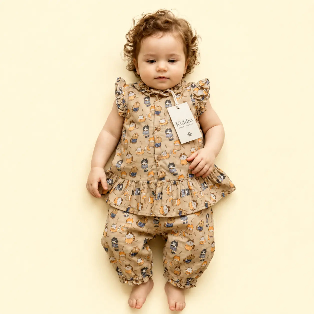 A toddler girl wearing a beige two-piece outfit with a cartoon animal print, featuring a ruffled sleeveless top and matching pants, lying on a solid pale yellow background.