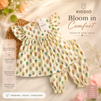 A baby girl's two-piece outfit set from Kiddio, featuring a cream-colored top and matching pants covered in a colorful floral pattern, displayed on a wicker basket with promotional text overlays.