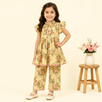 A young girl stands in a studio wearing a yellow floral two-piece outfit with flutter sleeves and matching pants. The set has a pink and green floral pattern on a pale yellow background with a pink ruffle trim.