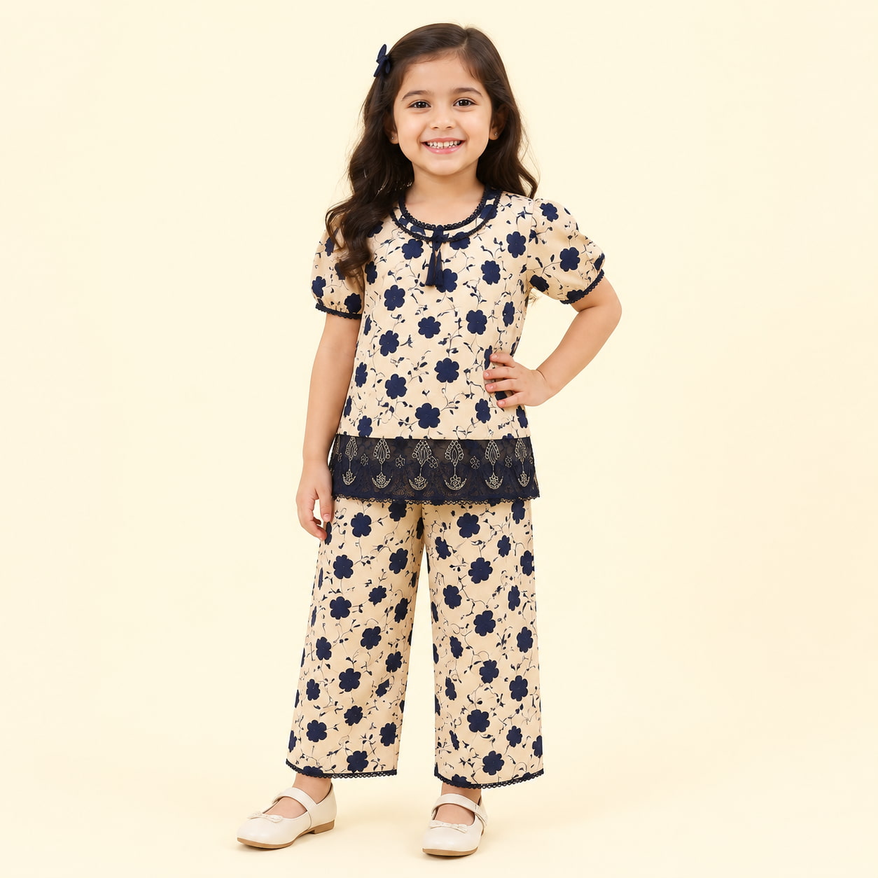 A young girl modeling a two-piece cream-colored pajama set with a navy blue floral pattern and lace trim, standing against a solid cream background.