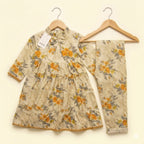 A two-piece children's outfit set consisting of a long-sleeve tiered dress and matching pants, both featuring a floral print in orange, yellow, and gray on a beige background. The dress has a ruffled collar with a bow, and both pieces are displayed on wooden hangers against a solid cream background.