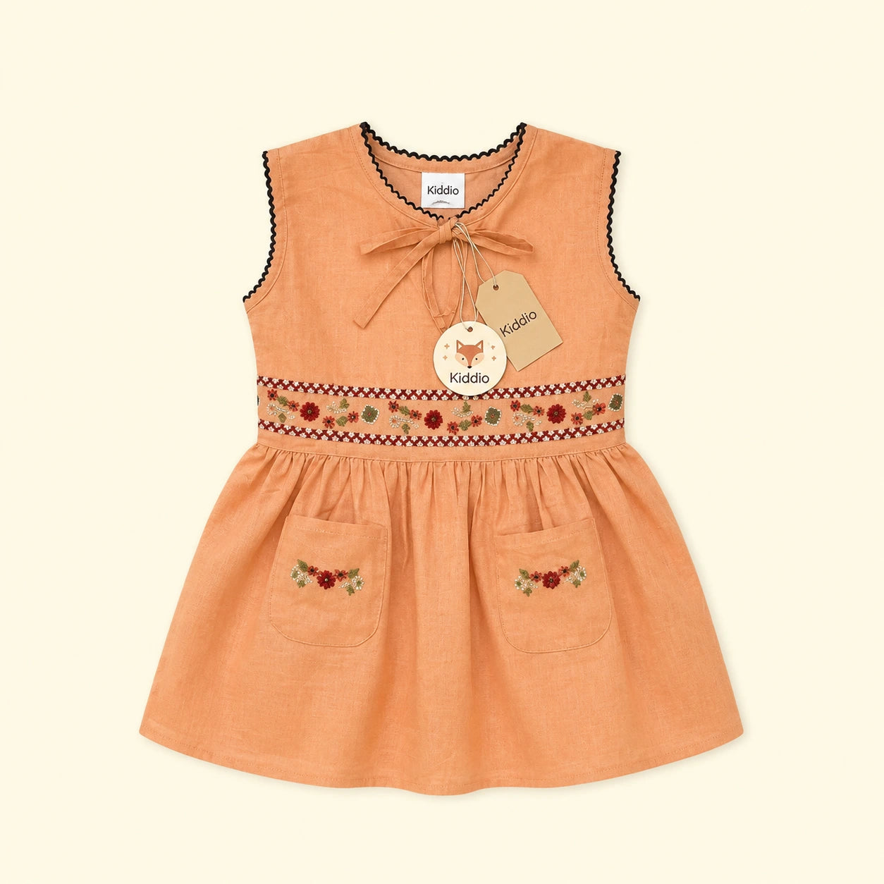 A sleeveless peach-colored toddler dress with black scalloped trim, a bow tie at the neckline, and embroidered details around the waist and on the front pockets. The dress features a paper hangtag with the brand name 'kiddo' visible on both the neck label and the hangtag.