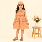 A young girl stands against a warm cream background wearing a peach sleeveless dress with embroidered details, a matching cap, and brown sandals. A small wooden stool with a vase of flowers is to her right.
