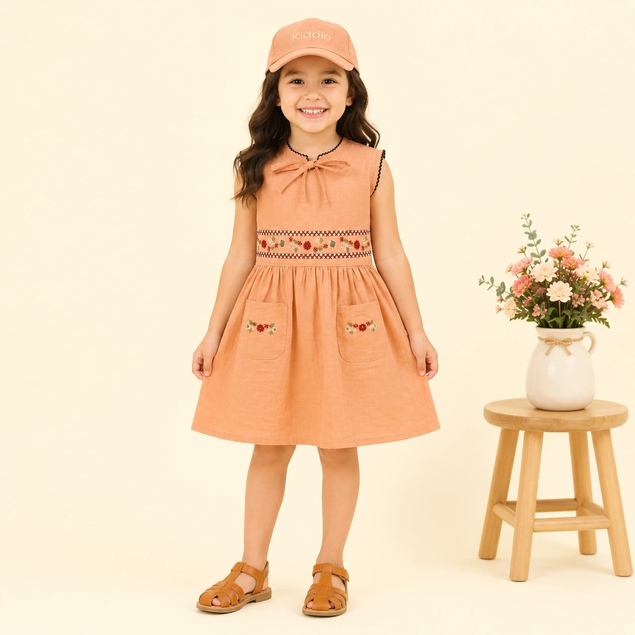 A young girl stands against a warm cream background wearing a peach sleeveless dress with embroidered details, a matching cap, and brown sandals. A small wooden stool with a vase of flowers is to her right.