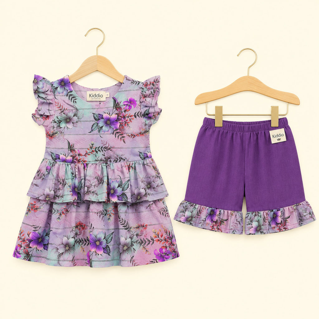 A toddler girl's two-piece outfit set consisting of a purple floral tiered ruffle dress and matching purple shorts with a floral ruffle hem, displayed on wooden hangers against a light cream background.