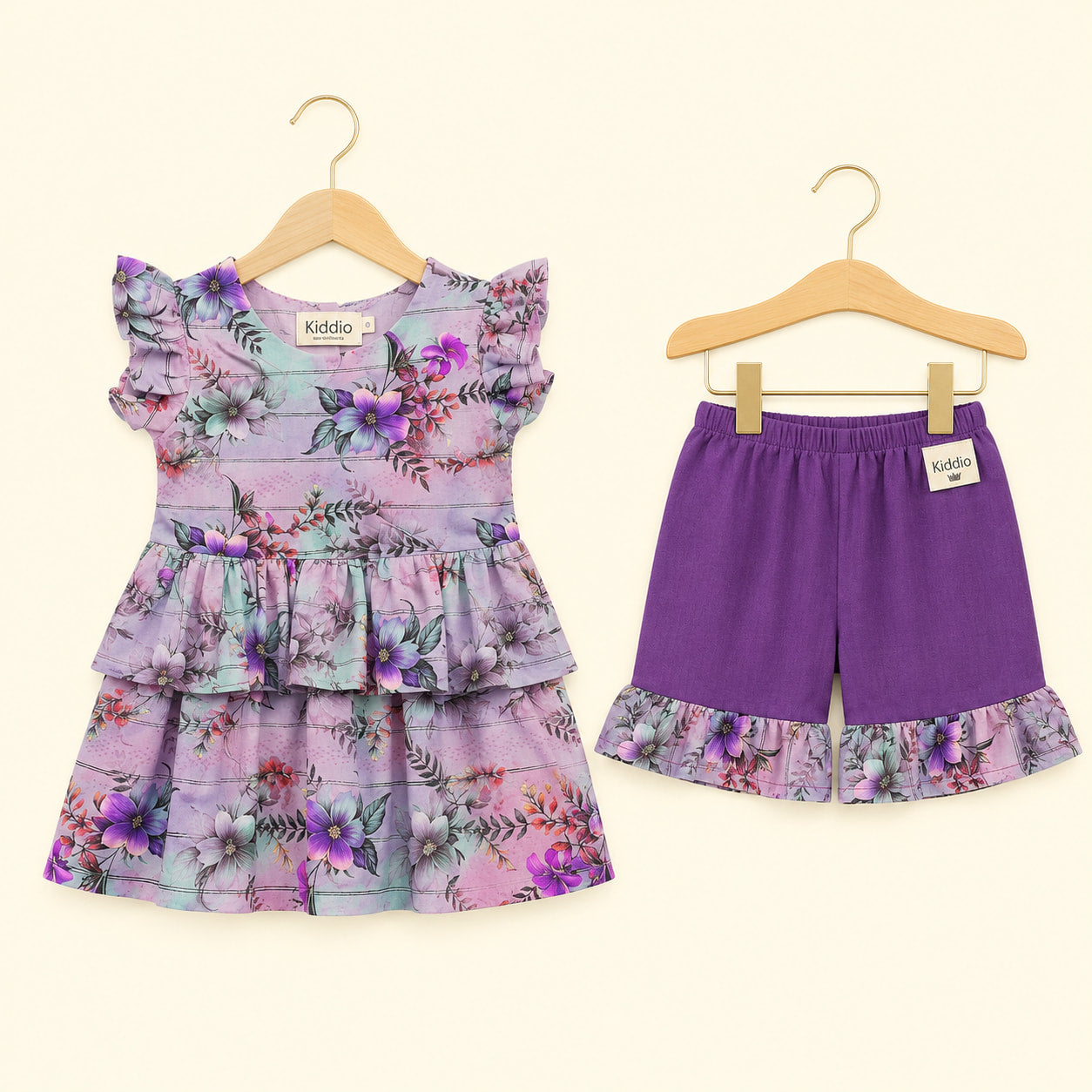 A toddler girl's two-piece outfit set consisting of a purple floral tiered ruffle dress and matching purple shorts with a floral ruffle hem, displayed on wooden hangers against a light cream background.