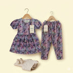 A baby clothing set from Kddio on wooden hangers against a pale cream background, featuring a short-sleeve dress, matching pants, and a small folded cream bodysuit. The dress and pants have a purple and lavender floral pattern.