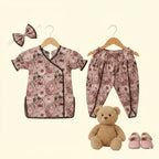 A baby clothing set displayed on hangers against a light cream background, including a short-sleeve floral top with dark trim, matching floral pants, a matching bow headband, a teddy bear, and pink shoes.