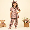 A young girl stands in a studio wearing a pink floral pajama set with black trim, a matching pink hat with a bow, and pink shoes. A teddy bear sits on the left, and a basket of flowers is on the right, against a warm cream background.