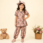 A young girl stands in a studio wearing a pink floral pajama set with black trim, a matching pink hat with a bow, and pink shoes. A teddy bear sits on the left, and a basket of flowers is on the right, against a warm cream background.