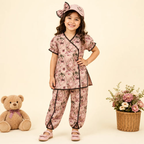 A young girl stands in a studio wearing a pink floral pajama set with black trim, a matching pink hat with a bow, and pink shoes. A teddy bear sits on the left, and a basket of flowers is on the right, against a warm cream background.