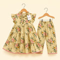A toddler girl's two-piece floral outfit set consisting of a tiered ruffle-sleeve dress and matching pants, hanging on wooden hangers against a light cream background. The set features a warm beige base with a pink, green, and yellow floral pattern.