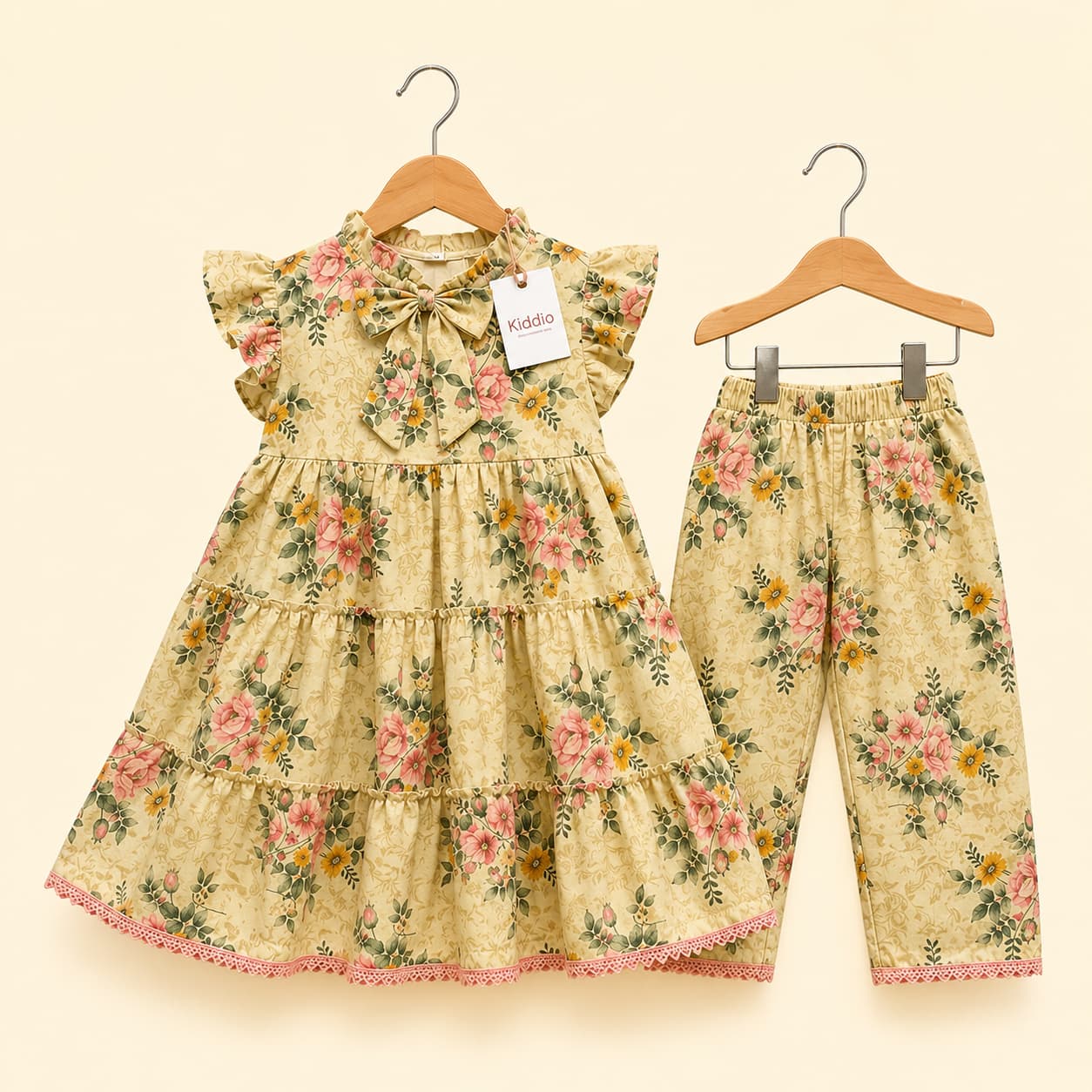 A toddler girl's two-piece floral outfit set consisting of a tiered ruffle-sleeve dress and matching pants, hanging on wooden hangers against a light cream background. The set features a warm beige base with a pink, green, and yellow floral pattern.