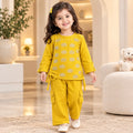 A young girl wearing a bright yellow two-piece outfit with beige circular patterns and white shoes, standing in a warm, modern living room.