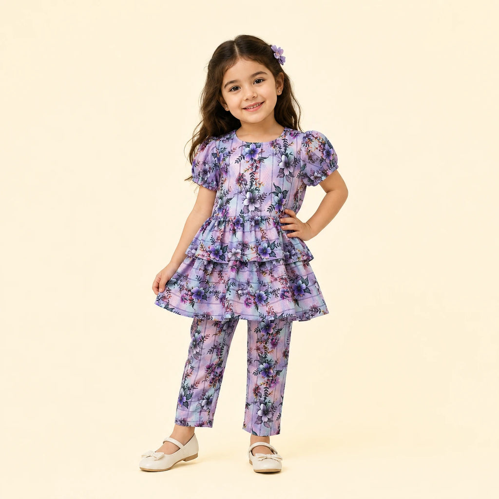 A young girl models a matching two-piece outfit consisting of a short-sleeve tiered tunic top and coordinating pants, both featuring a purple and blue floral pattern on a light lavender base. She is standing against a solid pale beige background.