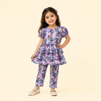 A young girl models a matching two-piece outfit consisting of a short-sleeve tiered tunic top and coordinating pants, both featuring a purple and blue floral pattern on a light lavender base. She is standing against a solid pale beige background.