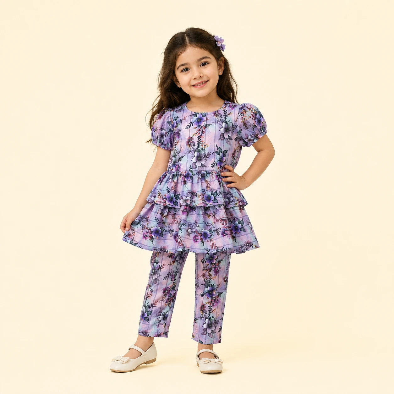 A young girl models a matching two-piece outfit consisting of a short-sleeve tiered tunic top and coordinating pants, both featuring a purple and blue floral pattern on a light lavender base. She is standing against a solid pale beige background.