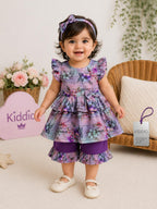 A toddler girl stands in a styled indoor studio setting wearing a purple floral tiered dress with matching headband and white shoes. The dress has a lavender base with purple, pink, and blue floral print, and solid purple ruffle trim.